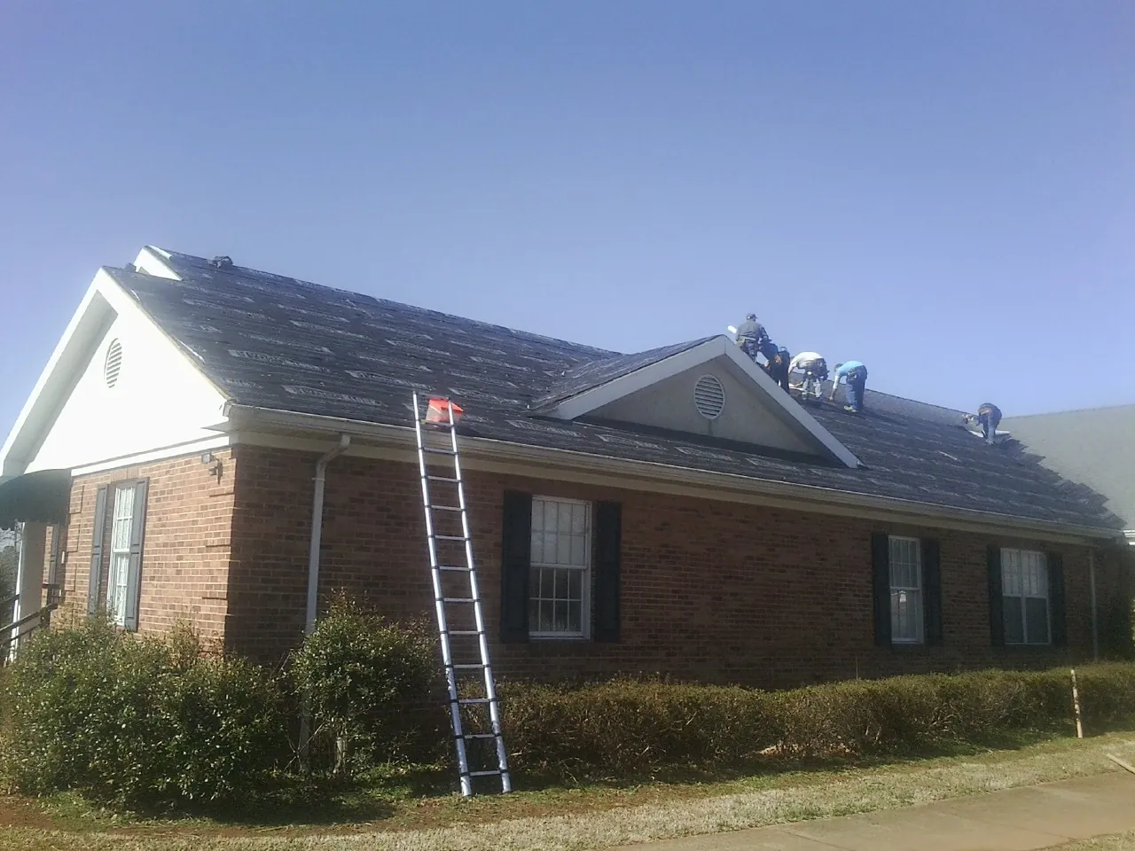 Crew tearing shingles off a brick ranch roof with a ladder set — Emergency Roof Repair work by our crew in Bumpass